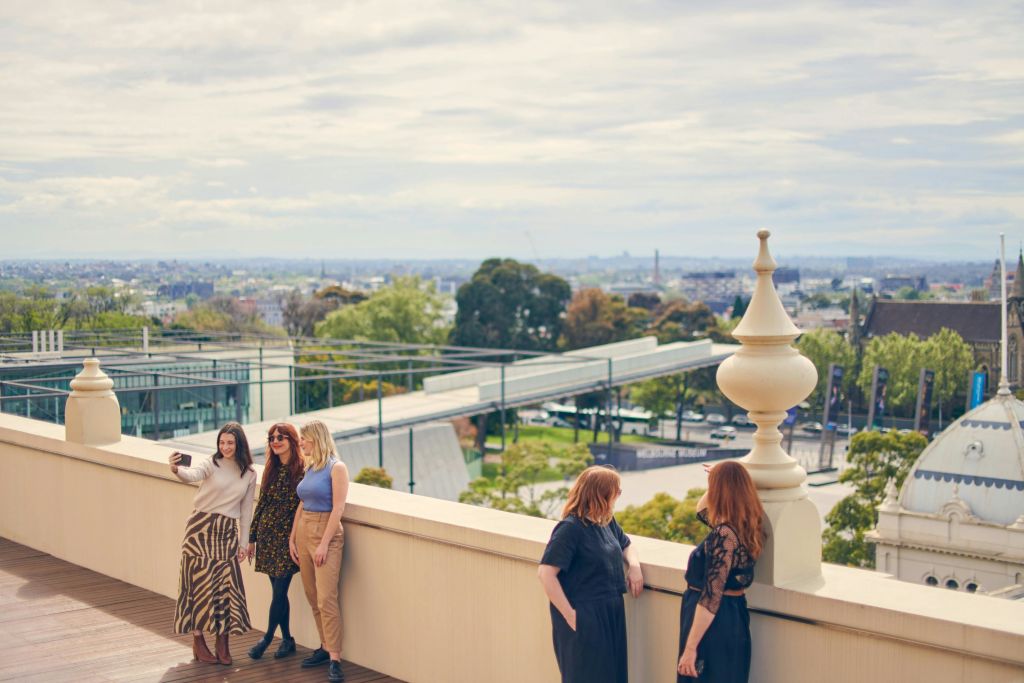 도시 풍경과 녹지를 배경으로 옥상에서 세 명의 여성이 셀카를 찍으며 포즈를 취하고 있으며, 근처에 또 다른 여성이 서 있습니다.