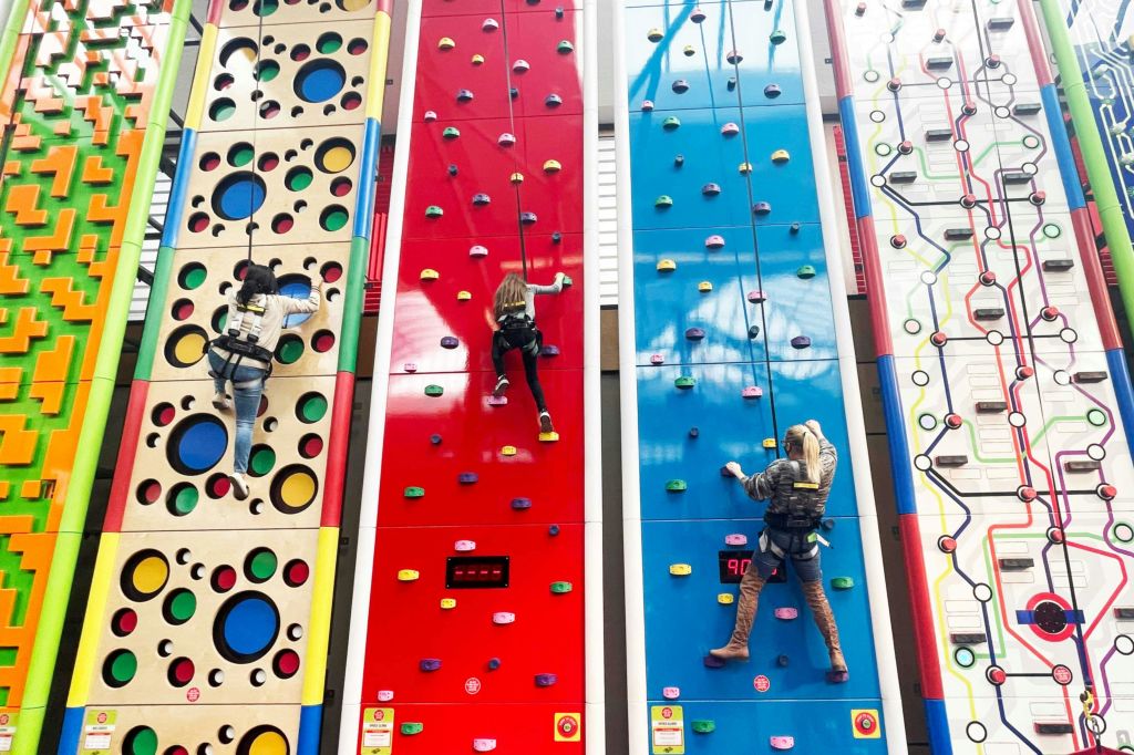 마구가 있는 다채로운 실내 등반 벽을 등반하는 세 사람, 각 벽에는 독특한 패턴과 그립이 있습니다.