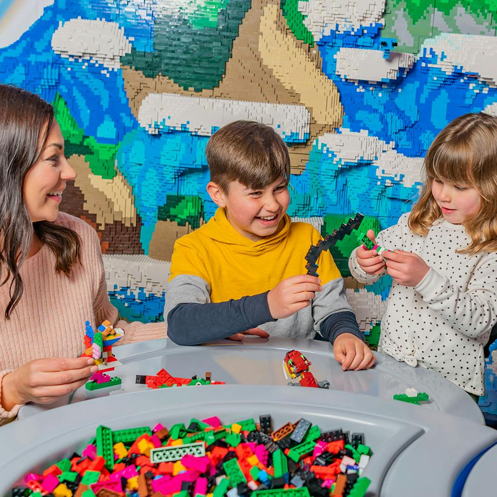 활기찬 풍경이 있는 레고로 지어진 벽면 앞, 테이블에서 화려한 레고 벽돌을 가지고 노는 세 명의 어린이.