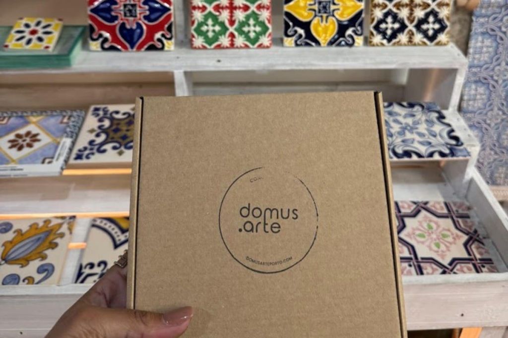 다채로운 장식 타일이 진열된 선반 앞에 "domus arte" 라고 적힌 판지 상자를 들고 있는 손.