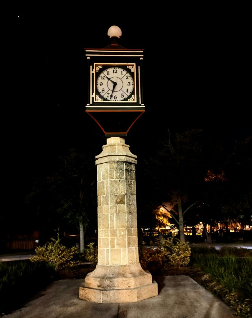 나무와 조명을 배경으로 어두운 야외 환경에서 조명을 받은 돌 받침대에 걸린 빈티지 시계입니다.