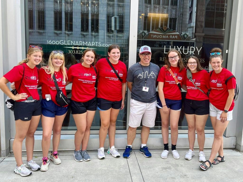 빨간색 셔츠를 입고 회색 셔츠를 입은 5명의 일행이 유리 상점 앞에 서 있습니다.