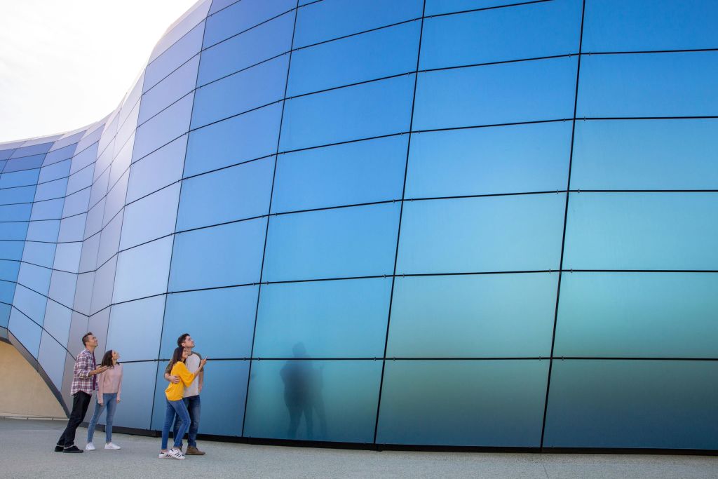 4명이 파란색 반사 패널이 있는 커다란 곡선형 건물을 올려다보고 있습니다.