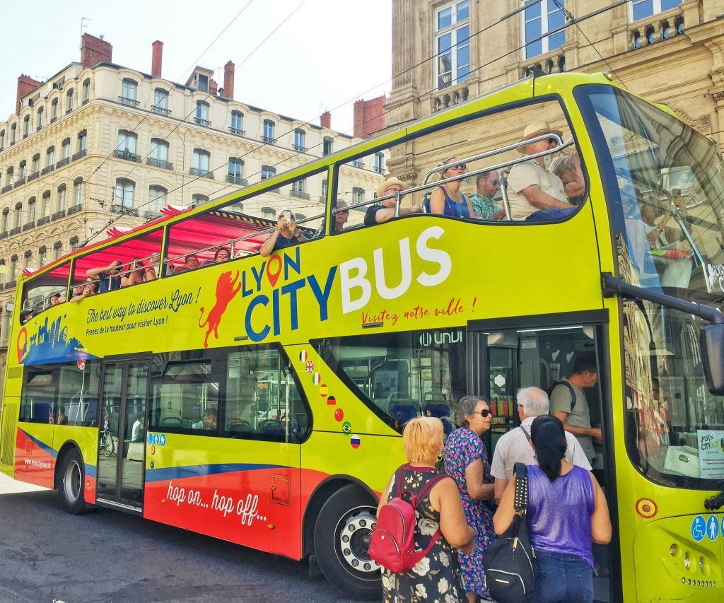 "Lyon City Bus" 라고 표시된 밝은 노란색과 빨간색의 2층 투어 버스가 도시에 주차되어 있습니다.