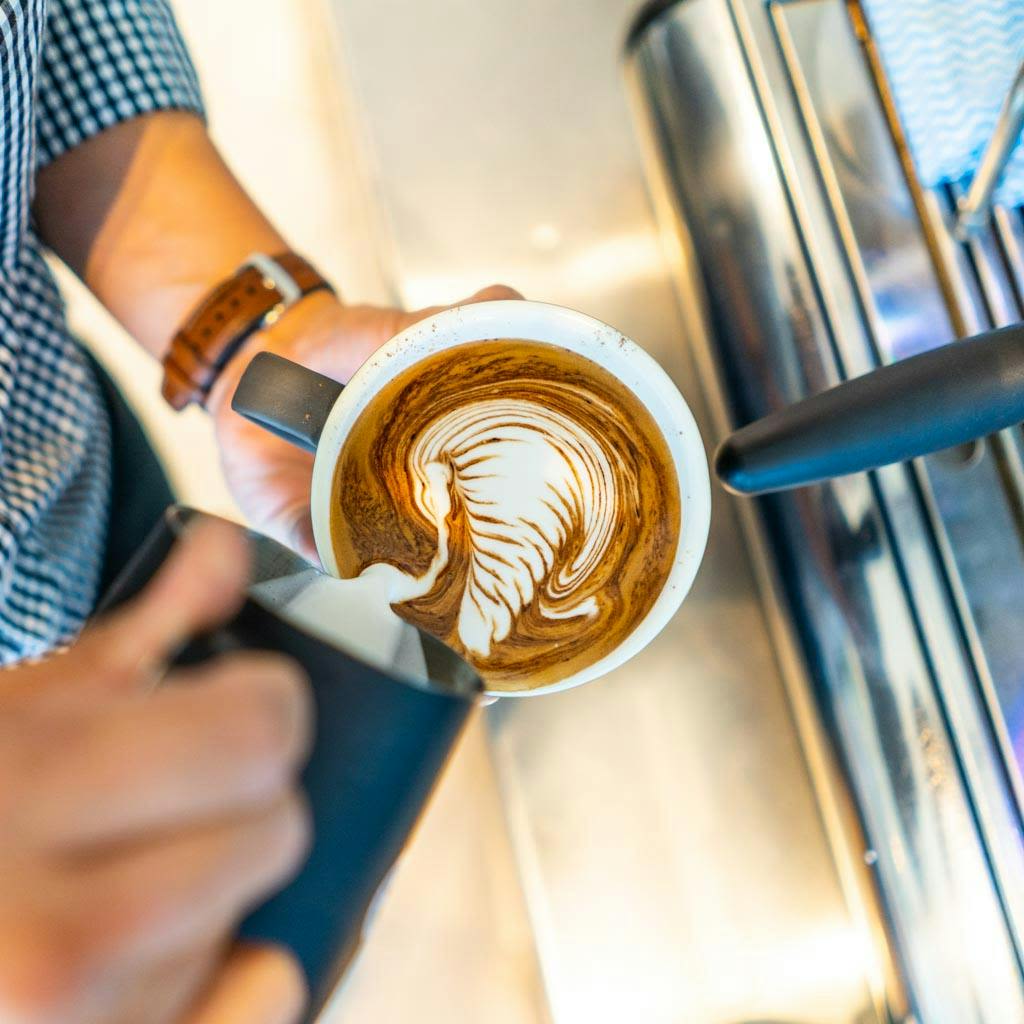 바리스타는 커피 한 잔에 찐 우유를 붓고 라떼 아트를 만듭니다. 오른쪽에 에스프레소 머신이 보입니다.