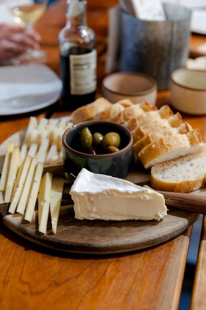 테이블 위에 얇게 썬 빵, 치즈, 올리브 한 그릇, 브리치즈 쐐기가 놓인 나무판.