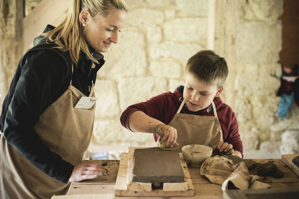 앞치마를 두른 한 여성과 한 소년이 소박한 분위기의 나무 테이블에서 점토로 작업하고 있습니다.