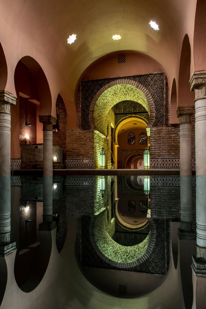 아치, 기둥, 벽돌 벽, 반사되는 수면이 있는 조명이 희미한 목욕탕의 내부.