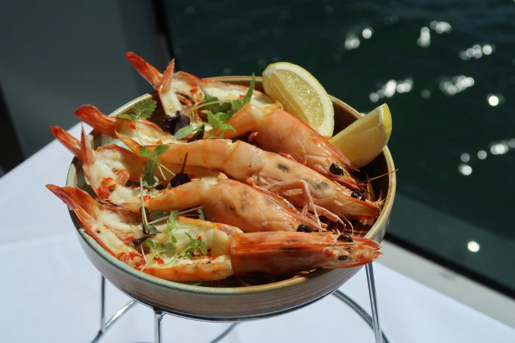 하얀 식탁보 위에 놓인 허브와 레몬 슬라이스로 장식된 구운 새우 그릇에 물이 배경으로 보입니다.