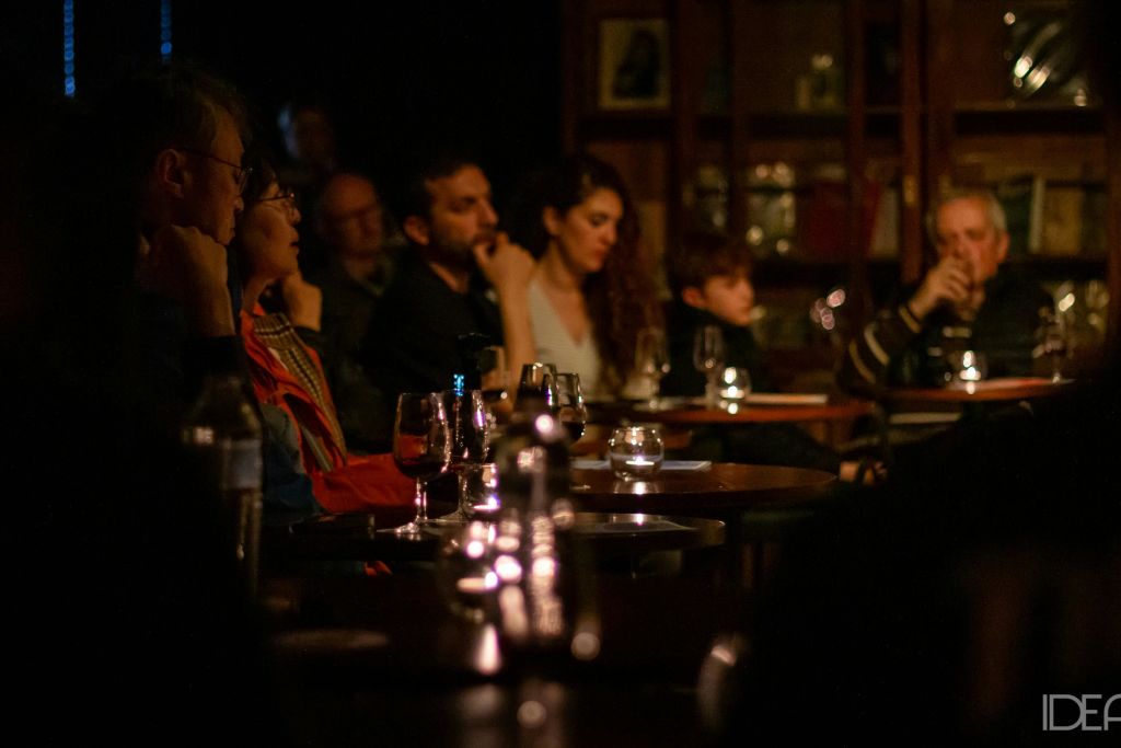 조명이 어두운 방에서 사람들이 테이블에 앉아 카메라 밖에서 무언가를 세심하게 관찰하고, 테이블 위에 안경과 촛불이 놓여 있습니다.