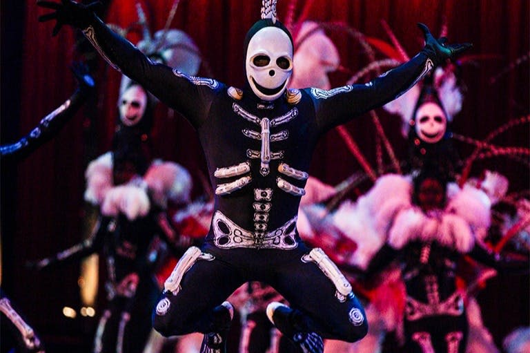 무대에서 춤을 추는 해골 의상을 입은 연주자. 비슷한 의상을 입은 연주자들과 붉은 커튼을 배경으로 춤을 추고 있다.
