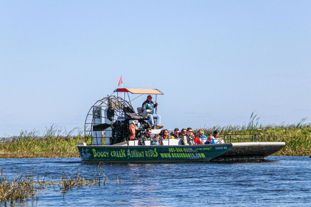 승객이 여러 명인 에어보트가 수로를 미끄러지듯 지나가고, 한 사람이 뒤에서 조종하고 있습니다. 잔디와 탁 트인 하늘이 배경입니다.