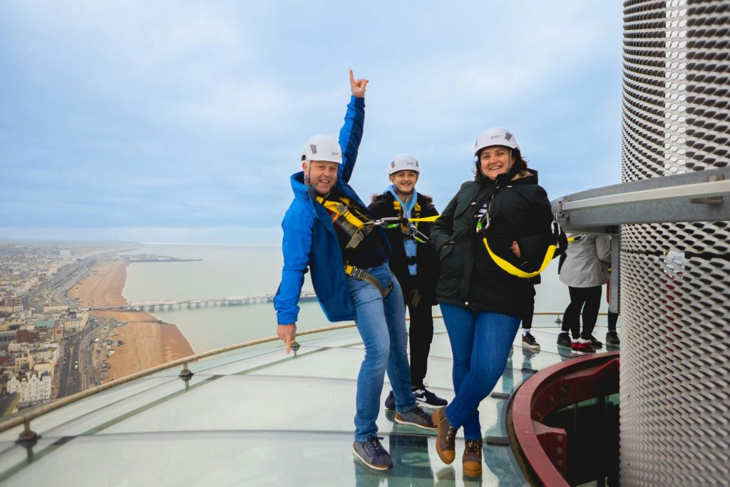 해안 전망을 배경으로 유리 전망대에 안전 장치와 헬멧을 착용한 세 사람이 서 있습니다.