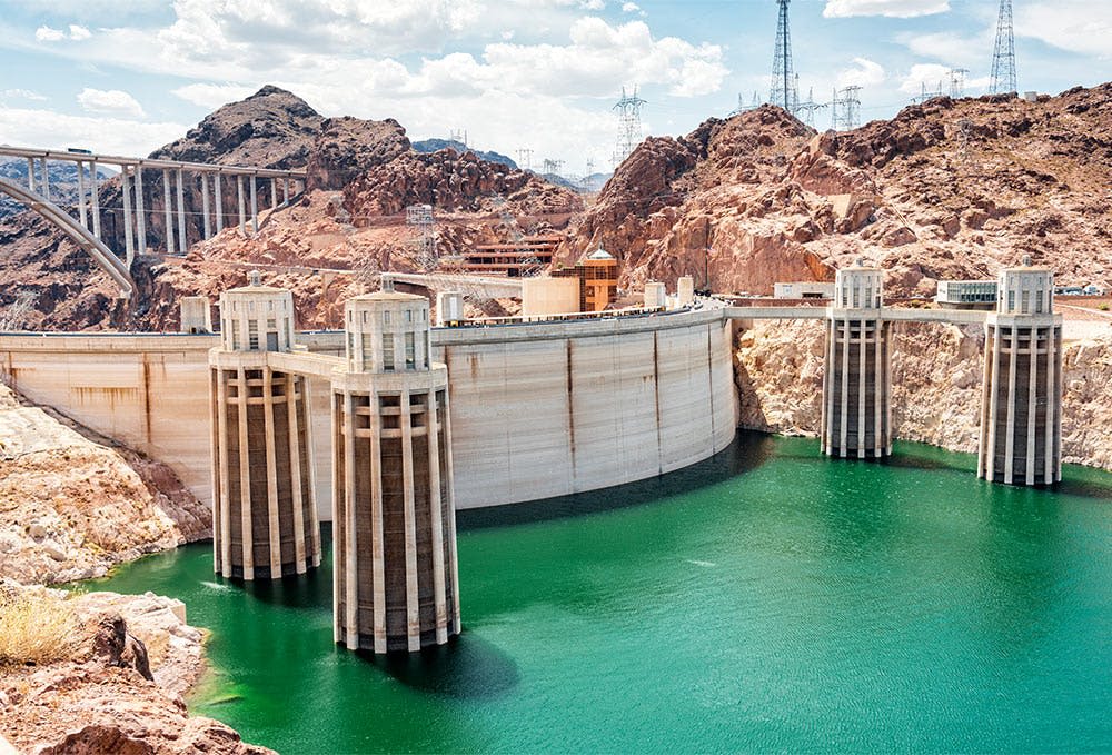 탑이 있는 커다란 댐은 전기 인프라와 푸른 하늘이 있는 바위 지형으로 둘러싸인 녹색 저수지에 걸쳐 있습니다.
