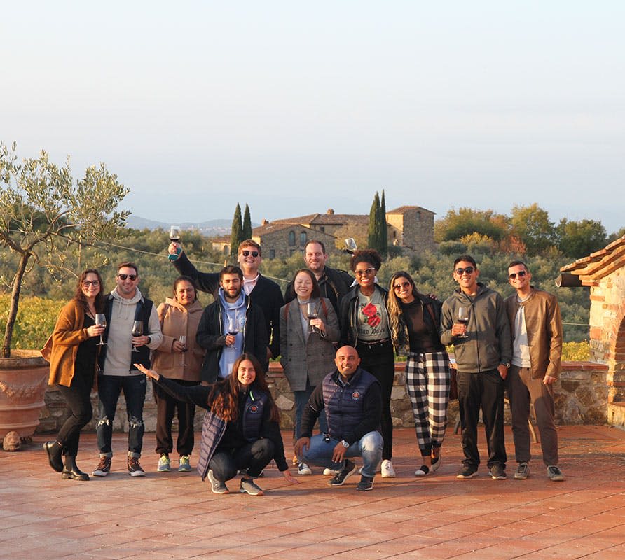 음료를 손에 들고 테라스에 서서 미소를 짓고 있는 12명의 일행, 경치 좋은 시골 풍경과 건물을 배경으로.