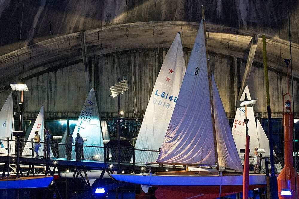 드라마틱한 조명이 있는 실내 전시물에 전시된 요트를 보는 사람들.