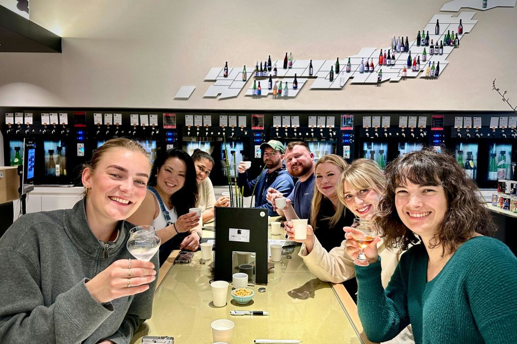 선반과 병을 배경으로 밝게 빛나는 방에서 7명이 미소를 지으며 테이블 주위에 컵을 올리고 있습니다.