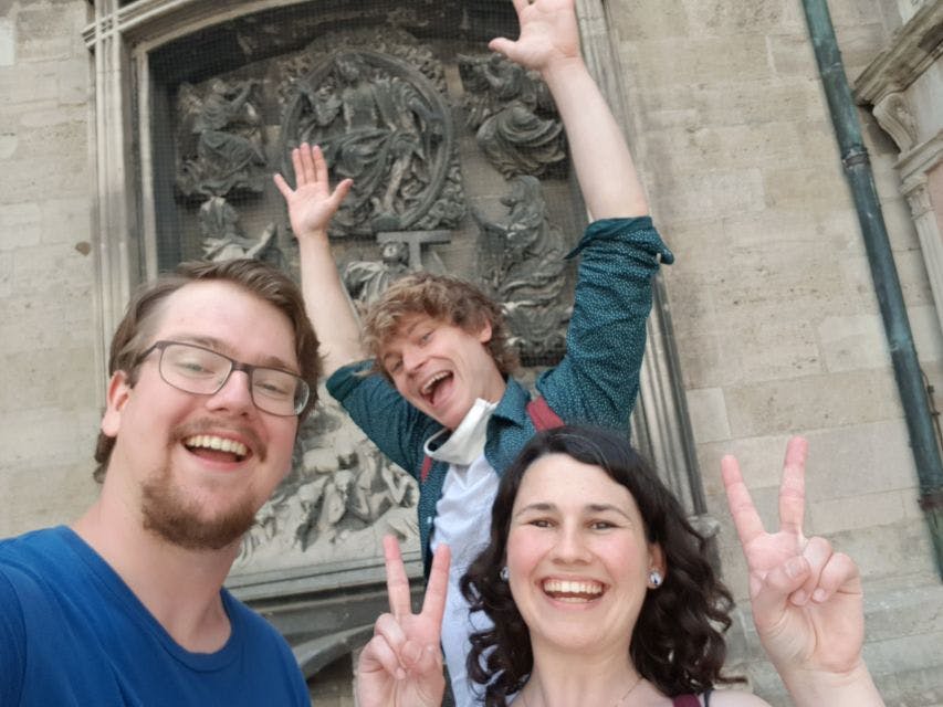 건물에 새겨진 석조 부조 앞에서 세 사람이 미소를 지으며 평화 표지판을 만들고 있습니다.