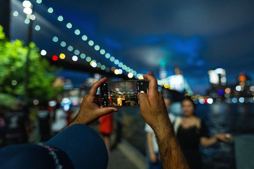 한 사람이 도시의 불빛을 배경으로 밤에 물가에서 포즈를 취하는 다른 두 사람의 모습을 스마트폰에 담아 사진을 찍고 있습니다.