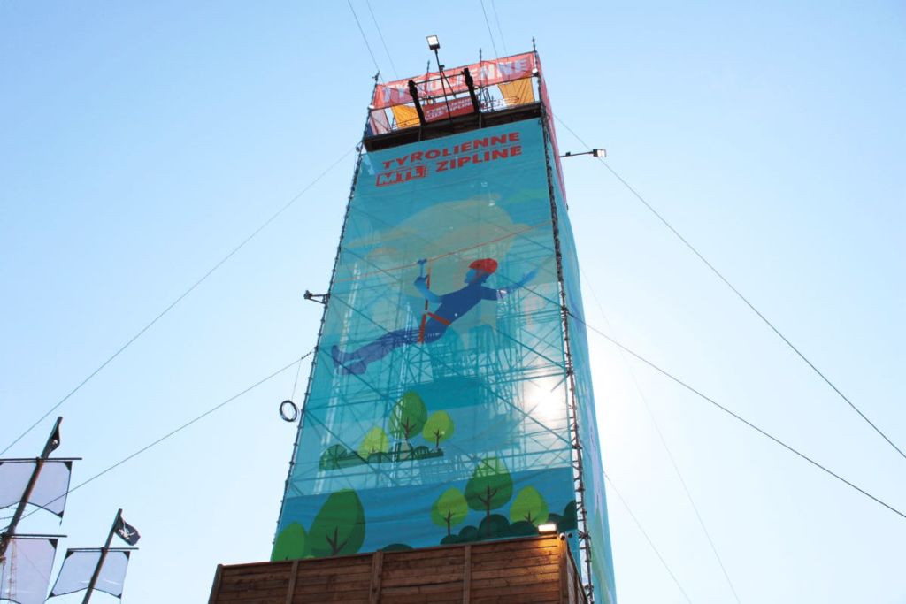 짚라인 설정이 가능한 높은 구조입니다. 표지판에는 'Tyrolienne/Zipline' 이라고 쓰여 있으며, 지퍼를 달고 있는 사람의 그림이 있습니다.