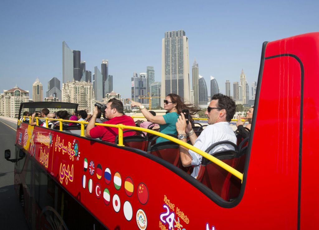 현대적인 고층 빌딩을 배경으로 도시의 스카이라인을 배경으로 빨간색 2층 버스의 상단 데크에서 사진을 찍는 관광객들.