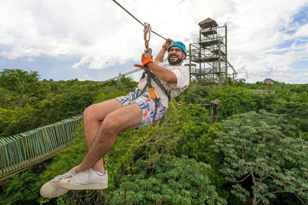 푸른색 헬멧과 화려한 색상의 반바지를 입은 사람이 높은 복층 전망대를 배경으로 무성한 녹지 위로 집라인을 타고 있습니다.