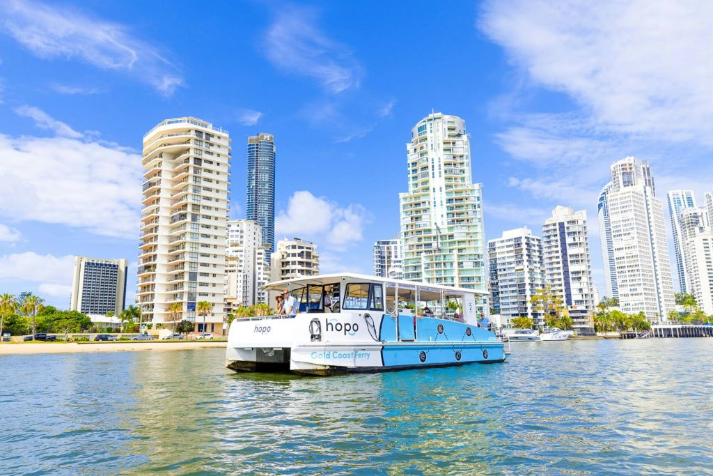 물가에 '호포 골드 코스트 페리 (Hopo Gold Coast Ferry)' 라는 이름의 페리가 있으며, 높은 건물이 햇살이 가득한 푸른 하늘을 배경으로 운행됩니다.
