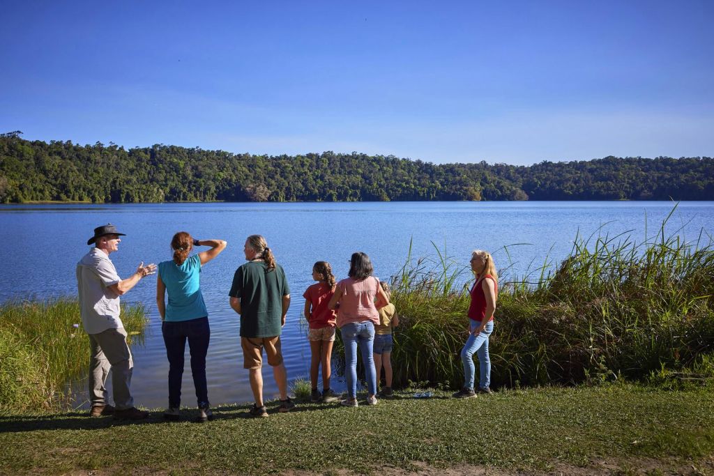 한 무리의 사람들이 푸른 하늘 아래 나무를 배경으로 잔디가 우거진 호숫가에 서 있습니다.