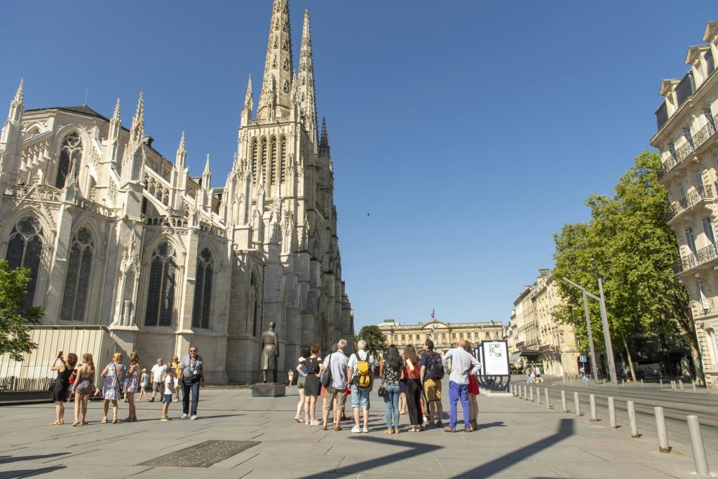 맑은 날 높이 솟은 첨탑이 있는 커다란 대성당 근처에 건물과 나무를 배경으로 한 무리의 사람들이 서 있습니다.