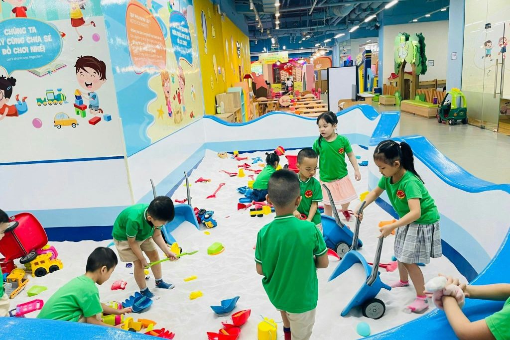 녹색 셔츠를 입은 아이들은 다채로운 장식과 교육용 포스터로 둘러싸인 실내 모래밭에서 장난감을 가지고 놀고 있습니다.