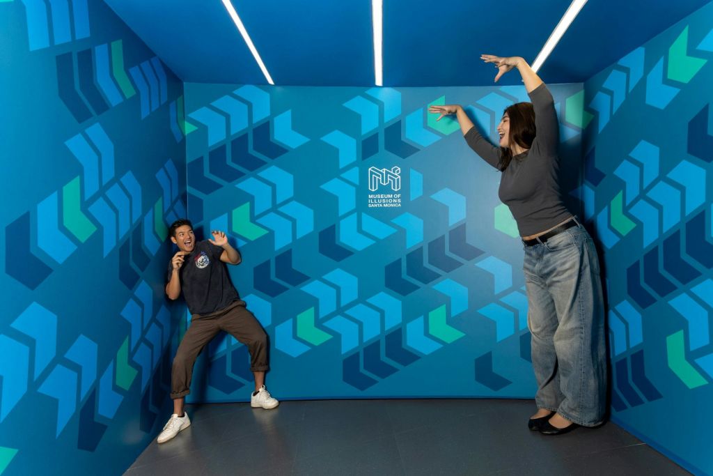 한 남성이 환상 박물관의 파란색 기하학적 무늬 벽을 배경으로 극적인 포즈를 취하고 있으며, 오른편에서 손을 뻗고 있습니다.