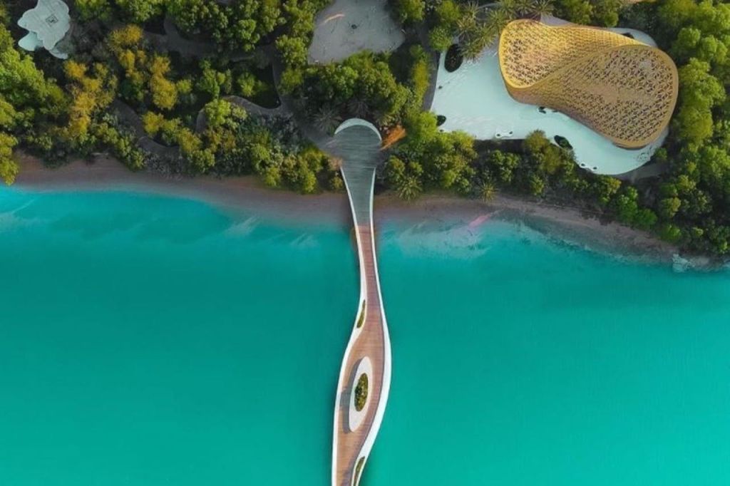 빽빽한 녹지와 현대적인 건축물로 둘러싸인 청록색 물 위로 뻗은 곡선형 보드워크의 공중 전망.