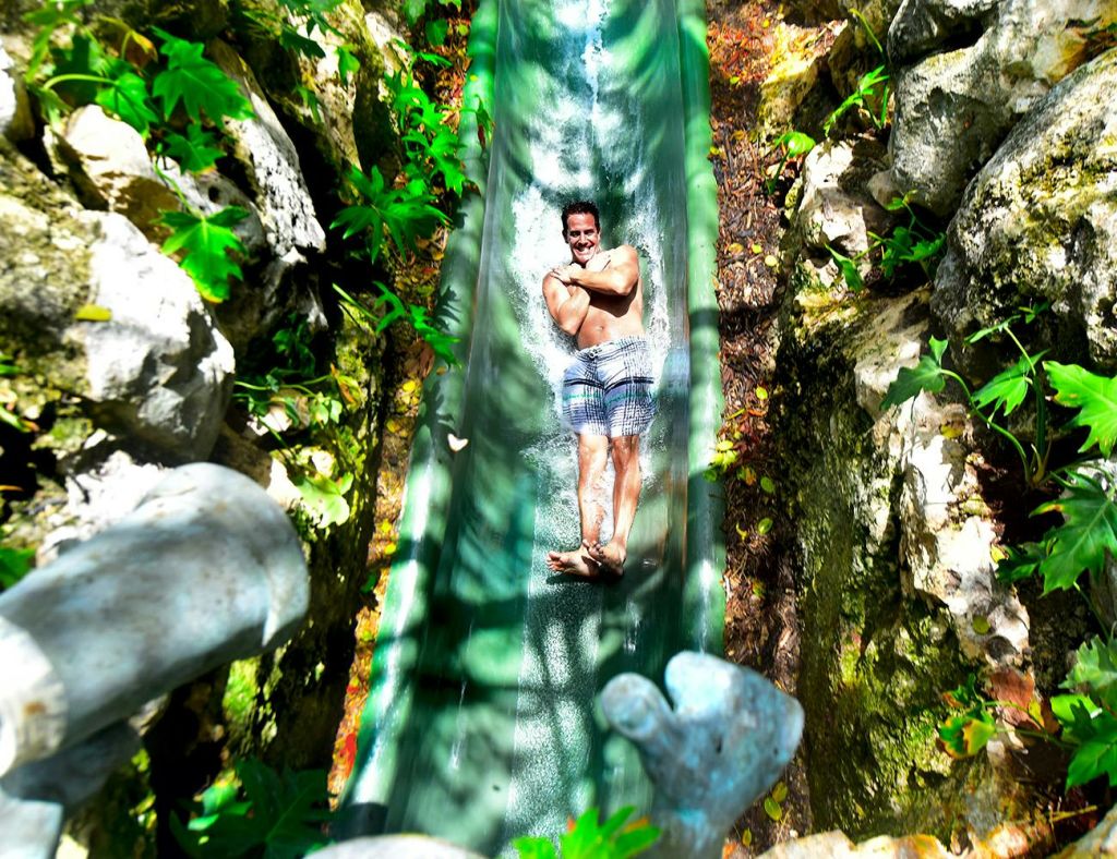 수영복을 입은 남성이 바위와 녹지로 둘러싸인 좁은 녹색 물 미끄럼틀을 미끄러지며 팔짱을 끼고 미소를 짓고 있습니다.