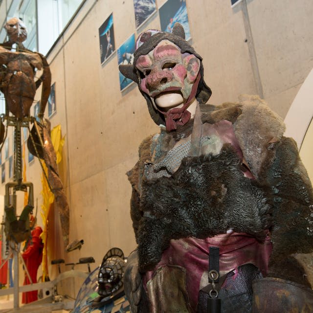 마스크와 모피를 포함한 괴물 같은 디테일한 의상을 입은 사람. 배경에는 조각과 콘크리트 벽이 포함됩니다.