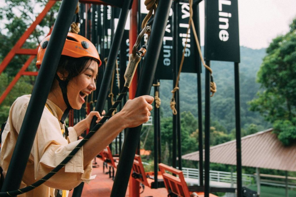나무를 배경으로 한 야외 어드벤처 코스에서 주황색 헬멧을 쓰고 미소를 짓고 밧줄을 잡고 있는 사람.