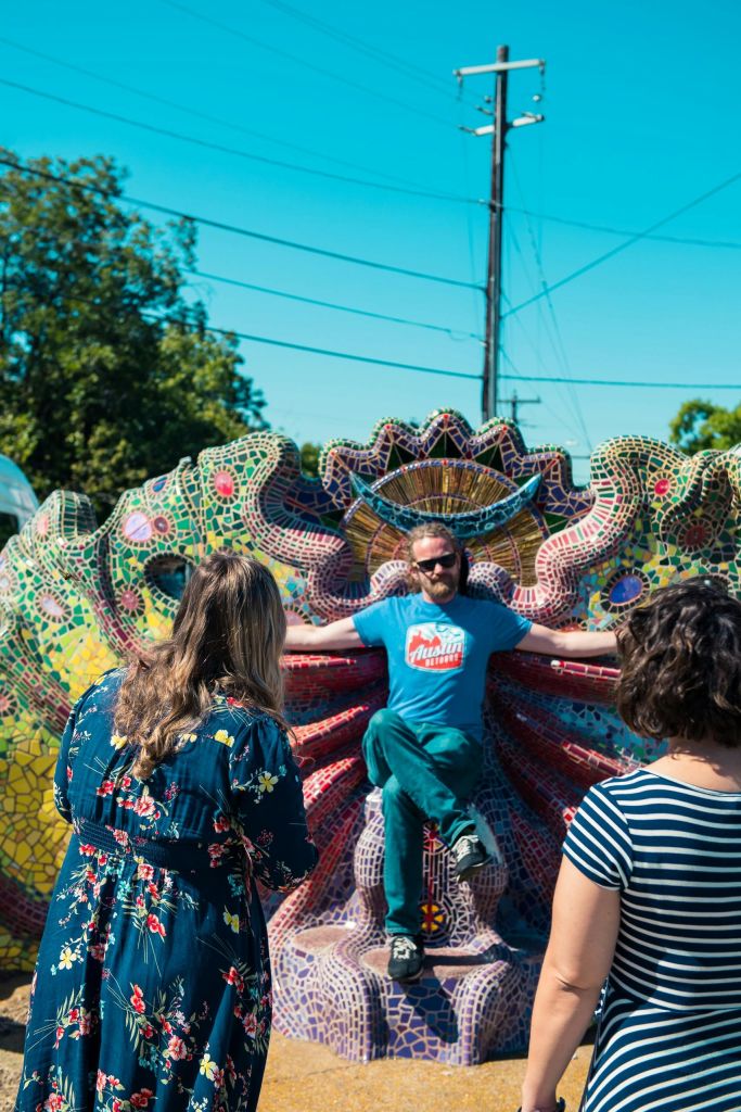 한 남자가 맑은 하늘 아래 야외에서 자신을 관찰하는 세 사람에 둘러싸인 화려한 모자이크 조각 위에 앉아 있습니다.