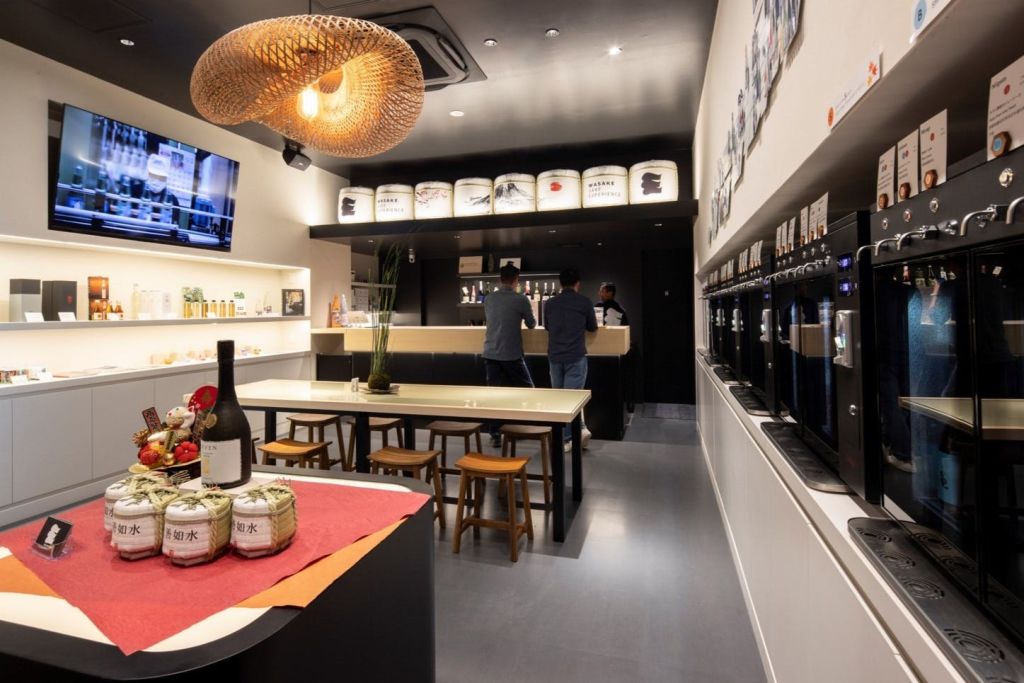 세 사람이 서 있는 모던한 바 인테리어, 스툴이 있는 긴 테이블, 사케 병, 벽에 일렬로 놓인 디스펜서.