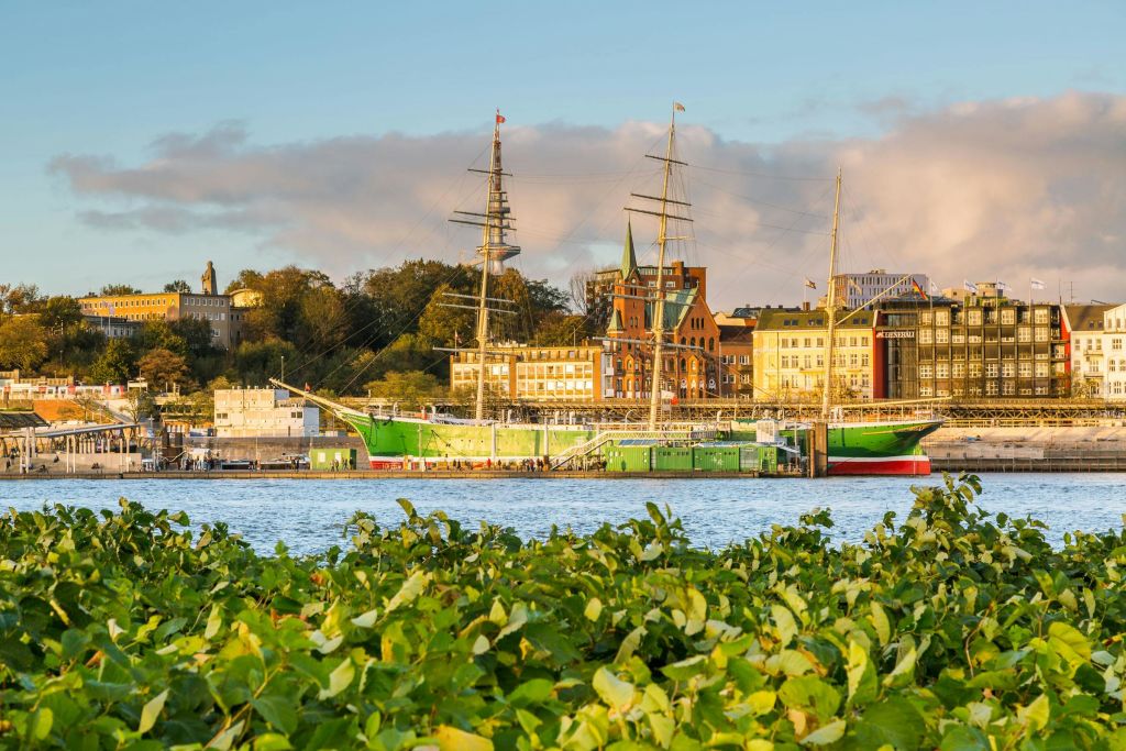 유서 깊은 건물이 늘어선 바닷가에 고층 선박이 정박해 있으며, 앞쪽에는 푸른 단풍이, 위쪽에는 맑은 하늘이 펼쳐져 있습니다.