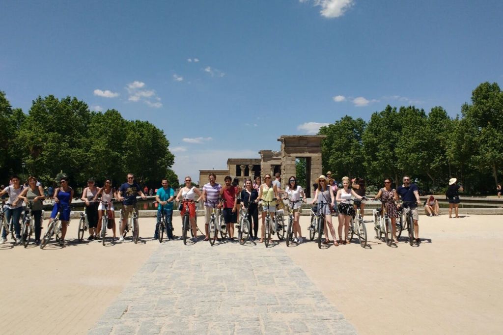 화창한 날 나무로 둘러싸인 고대 석조 건물 앞에 자전거를 탄 사람들이 서 있습니다.