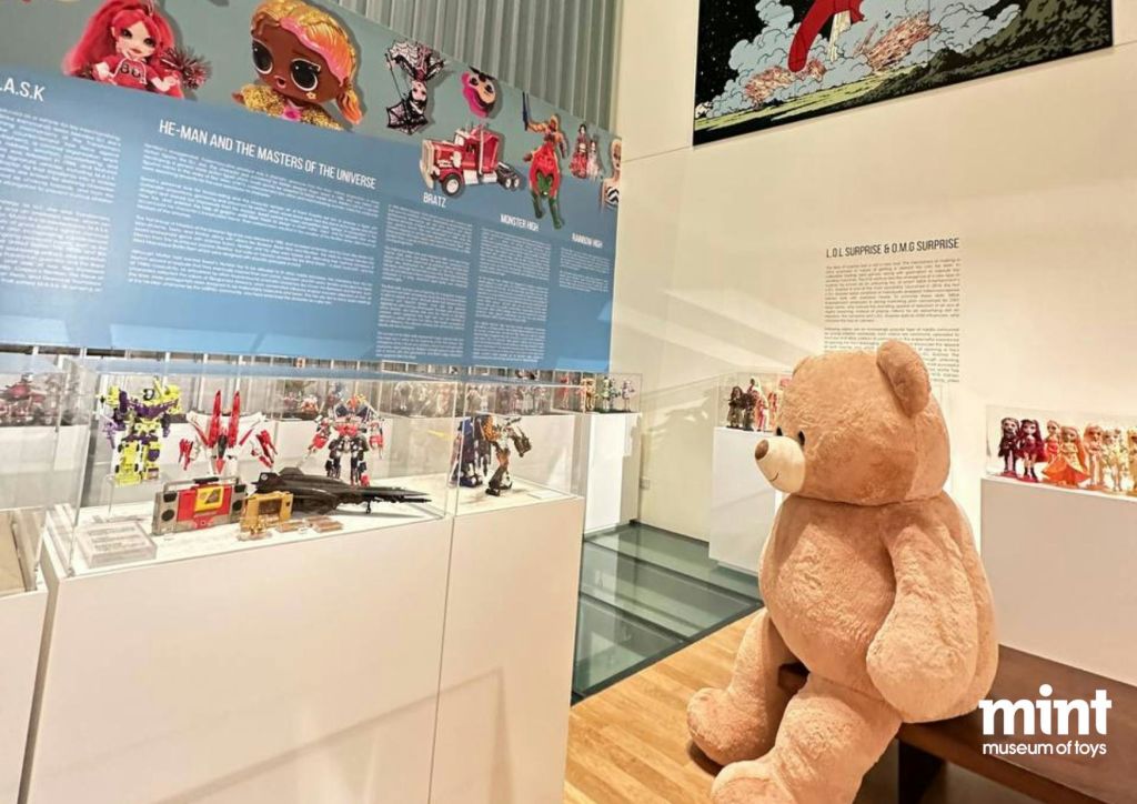다양한 액션 피규어와 장난감을 전시하는 박물관 전시관으로, 대형 테디베어가 전면에 배치되어 있습니다.