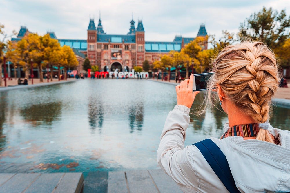 'I amsterdam' 표지판과 반사 수영장을 가로지르는 커다란 건물의 사진을 찍고 있는 사람으로, 양쪽에 나무가 있습니다.
