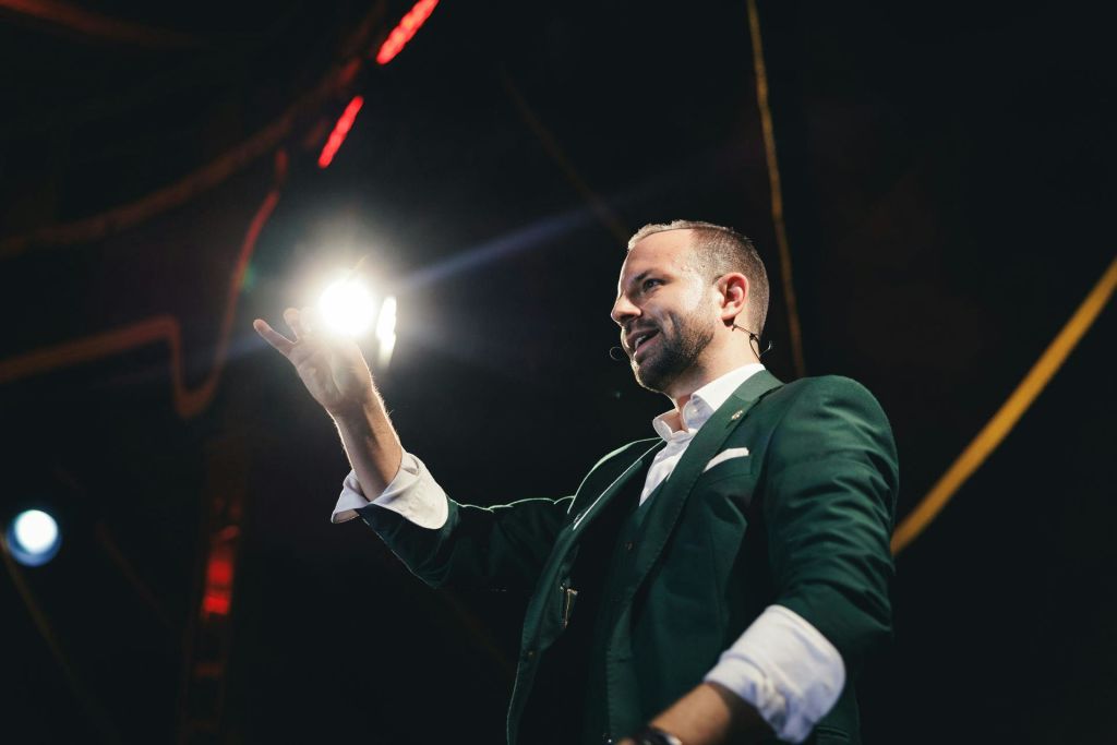 무대에서 마이크 헤드셋으로 말하고 어두운 환경에서 스포트라이트로 밝게 빛나는 녹색 블레이저를 입은 남성.