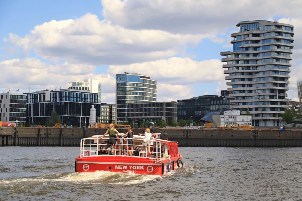 현대적인 건물과 부분적으로 흐린 하늘을 배경으로 선미에 "뉴욕 함부르크" 가 있는 작은 붉은 보트가 물 위를 여행합니다.
