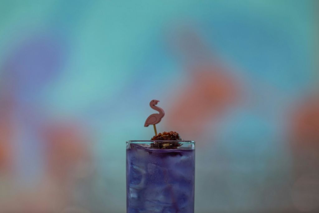 얼음이 담긴 보라색 음료 한 잔과 홍학 모양의 교반기가 흐릿한 다채로운 배경에 세팅되어 있습니다.