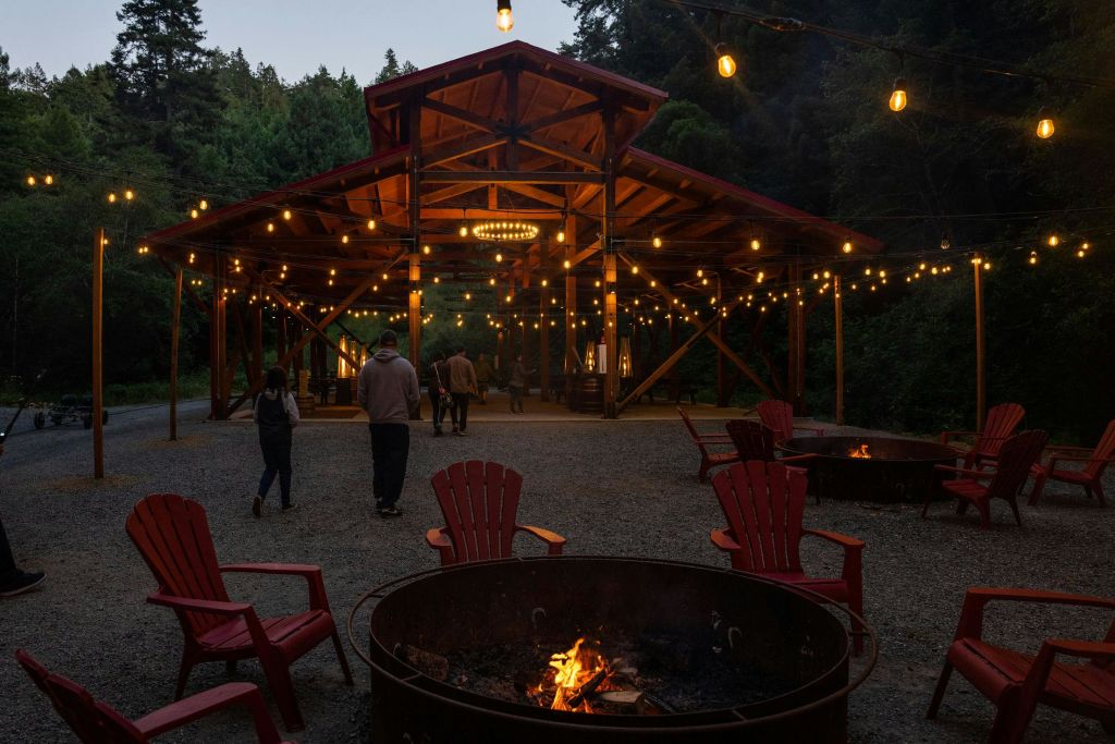 사람들은 황혼빛 하늘 아래 붉은 의자와 화덕으로 둘러싸인 매달린 조명이 있는 파빌리온을 향해 걸어갑니다.