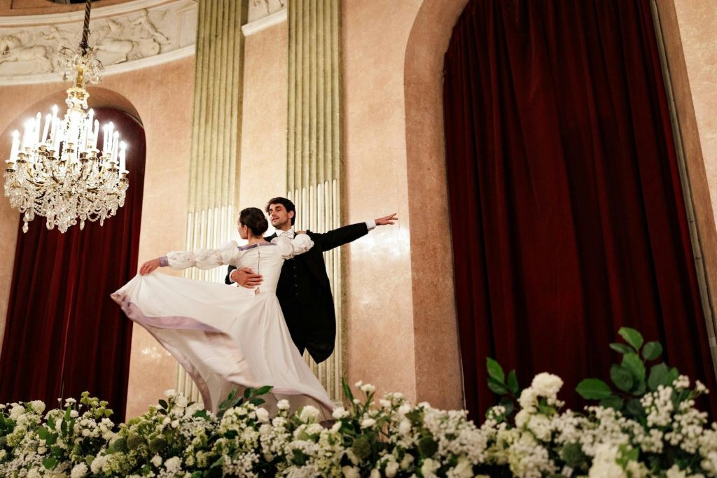 정장 차림의 커플이 무대 위에서 샹들리에, 빨간 커튼, 하얀 꽃을 배경으로 춤을 추고 있습니다.