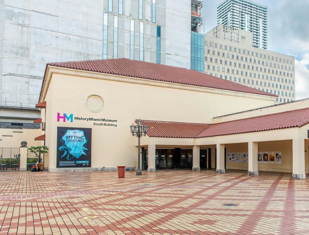 빨간색 타일로 지붕이 있는 마이애미 박물관 남쪽 건물 입구, "신화 속 생물" 전시 포스터, 고층 건물이 인접해 있습니다.