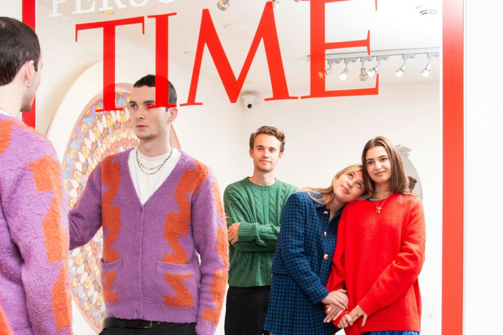 현대적인 인테리어가 돋보이는 밝은 방에서 화려한 의상을 입은 네 사람이 커다란 빨간색 텍스트 아래에서 포즈를 취하고 있습니다.