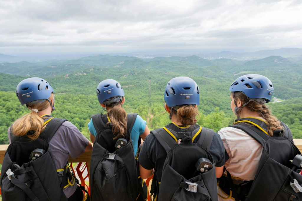 헬멧과 배낭을 착용한 세 사람이 플랫폼에 서서 흐린 하늘 아래 푸른 산이 우거진 풍경을 내려다보고 있습니다.