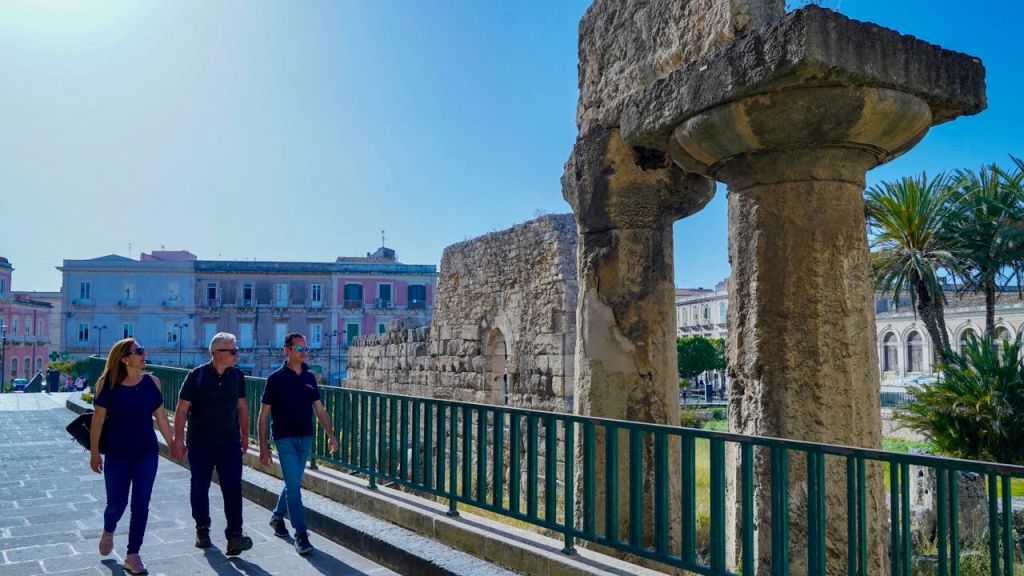 맑고 푸른 하늘 아래 고대 유적과 파스텔 색상의 건물 옆으로 돌로 포장된 길을 따라 세 사람이 걸어가고 있습니다.